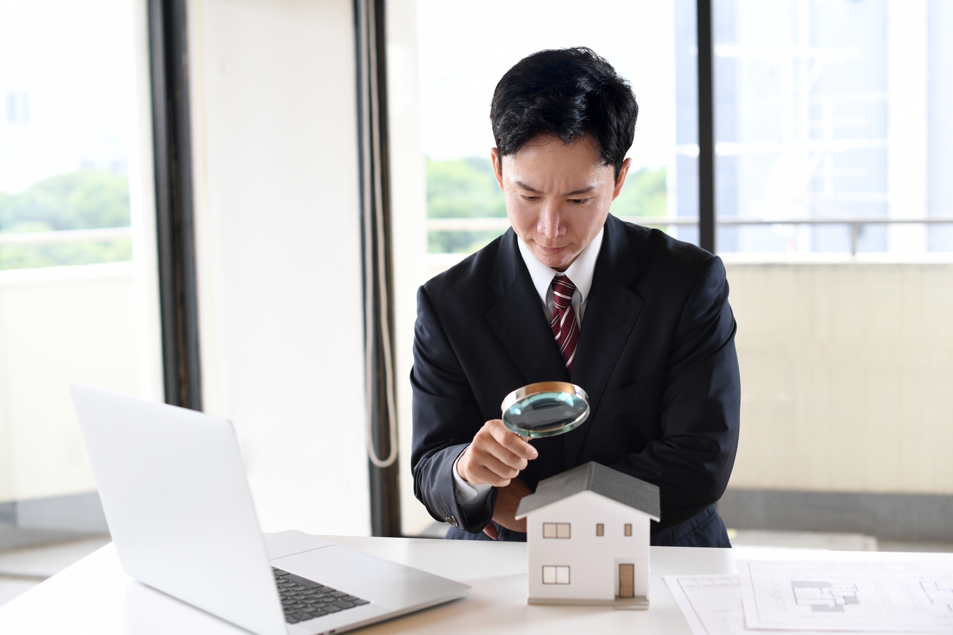虫眼鏡で住宅模型を覗くスーツ姿の若い男性