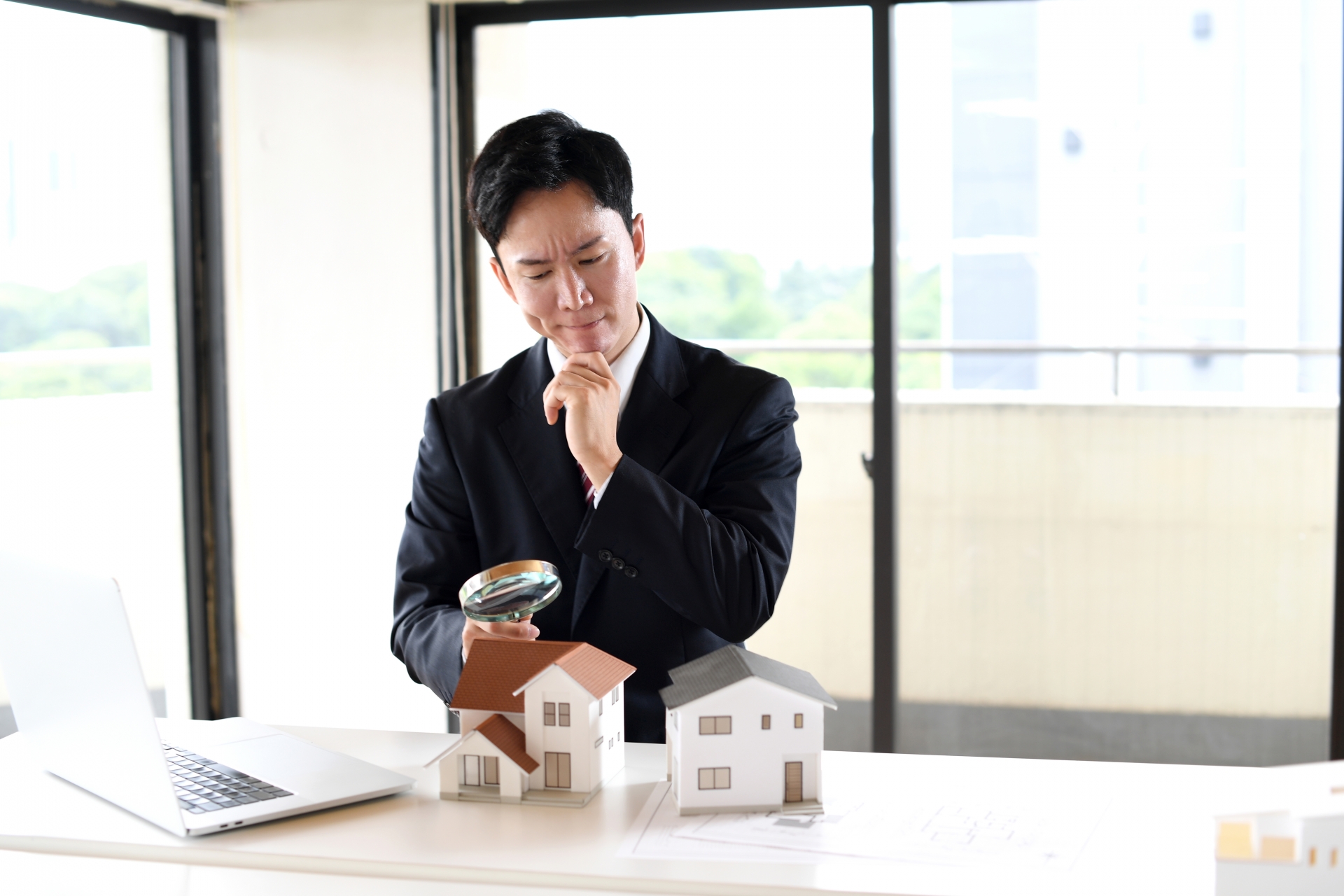 虫眼鏡で住宅模型を覗くスーツ姿の若い男性