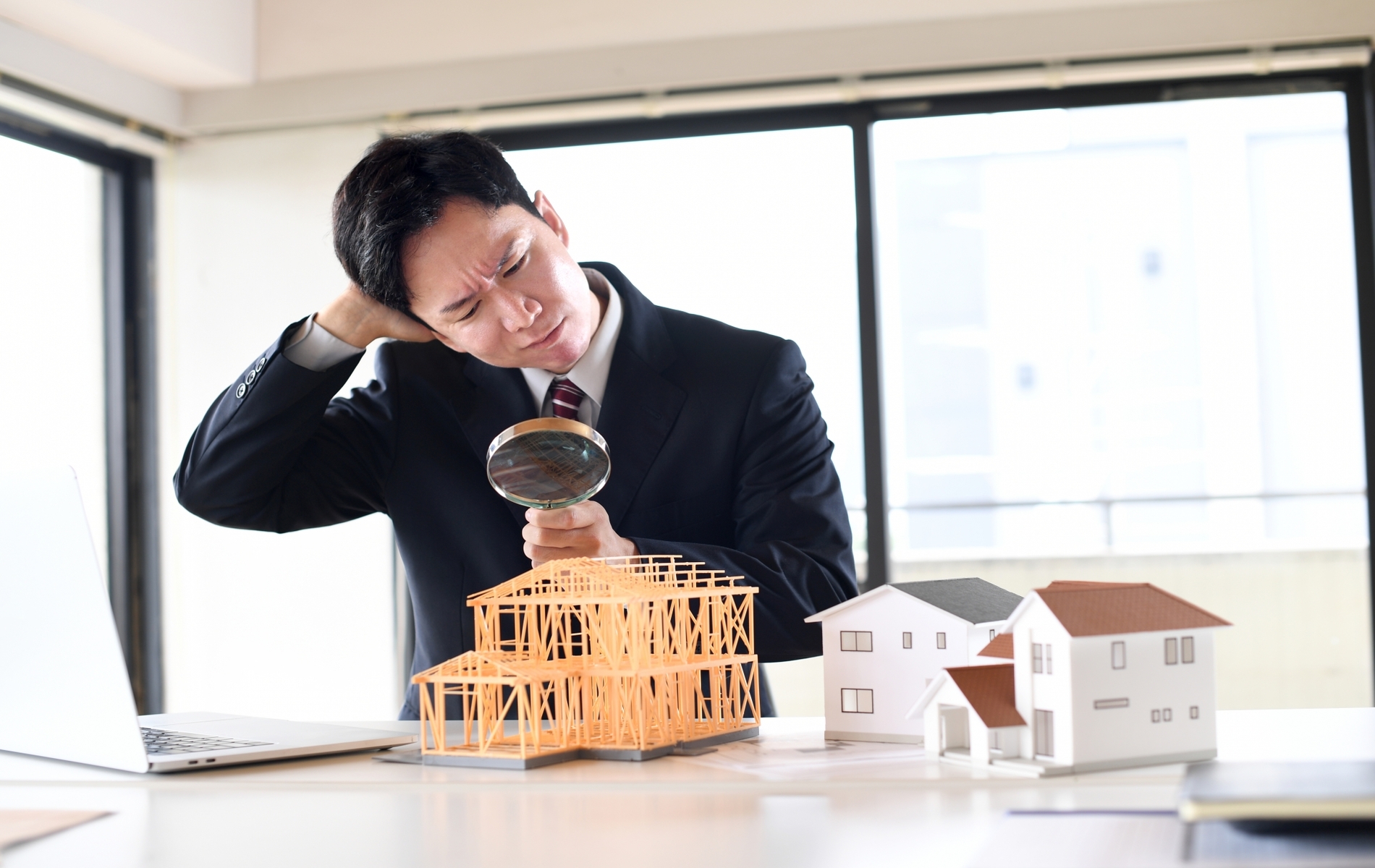 虫眼鏡で建築模型を覗くスーツ姿の悩む男性