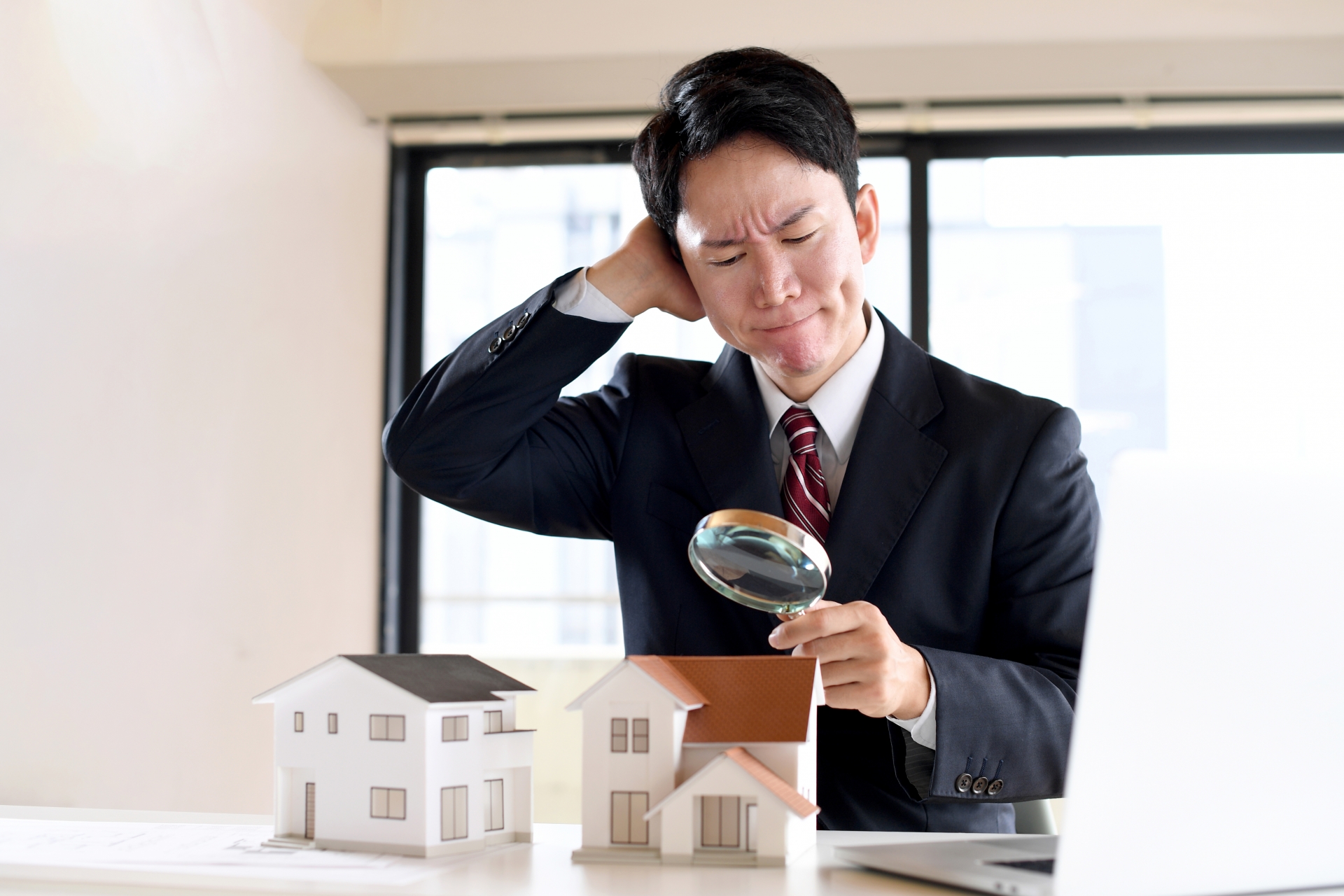 虫眼鏡で住宅模型を覗くスーツ姿の悩む男性